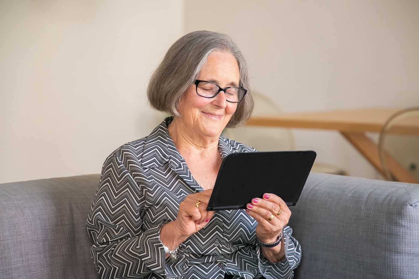 Senhora sorrindo enquanto lê no tablet, sentada no sofá de casa